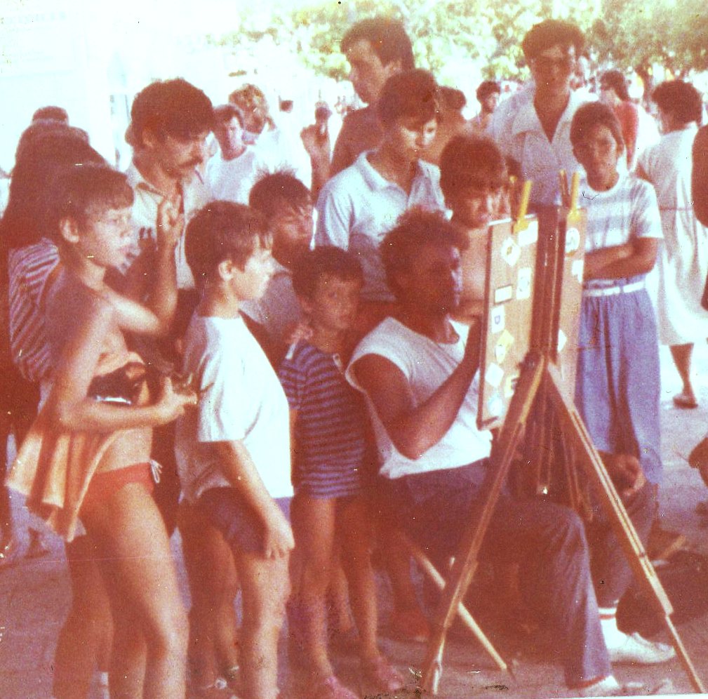 A crowd of children and adults gathered around Goran Goda as he draws at the easel, Baska Voda, 1985