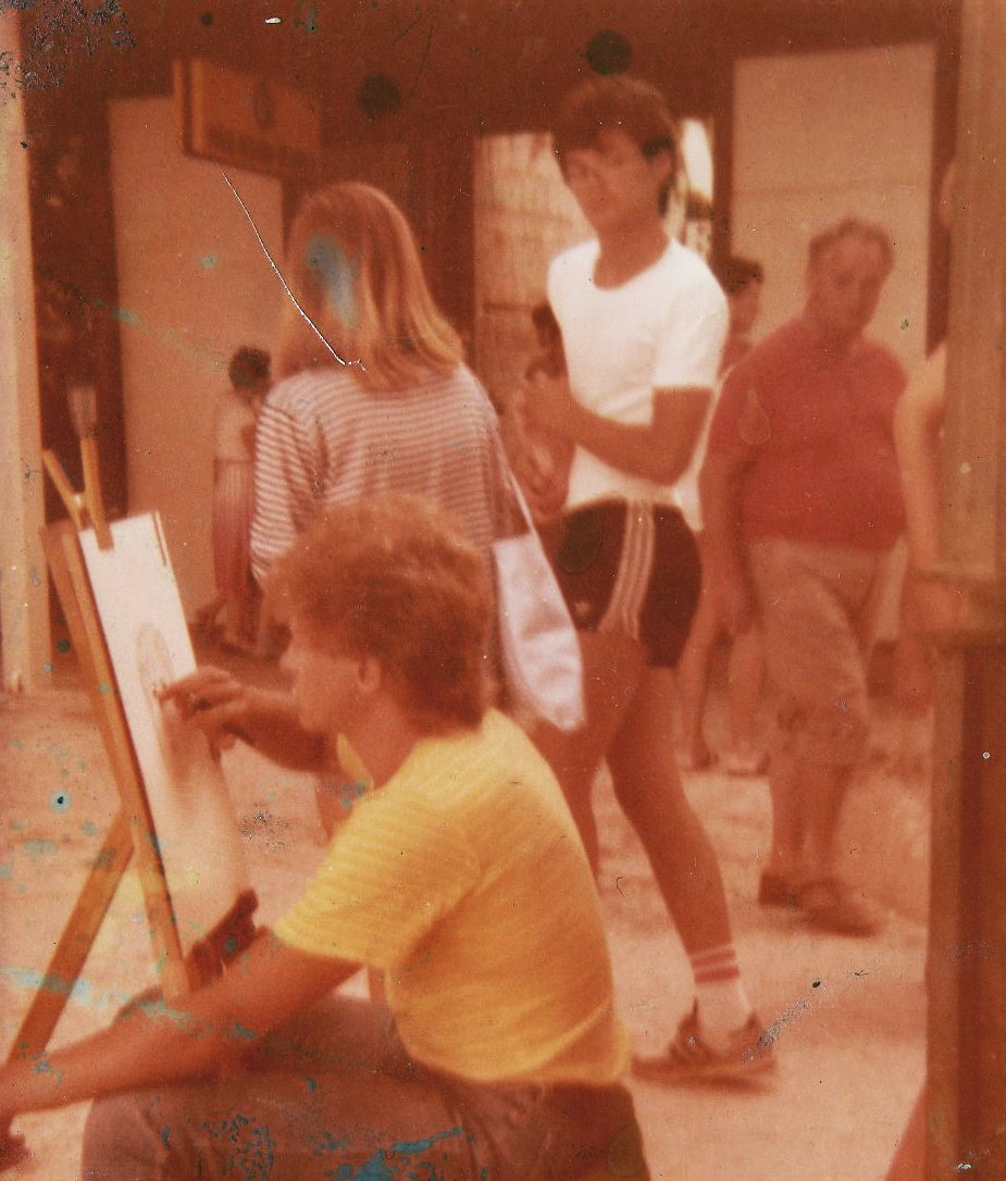 Goran Goda in a yellow shirt drawing a portrait at the easel while passersby watch, Brela, 1984